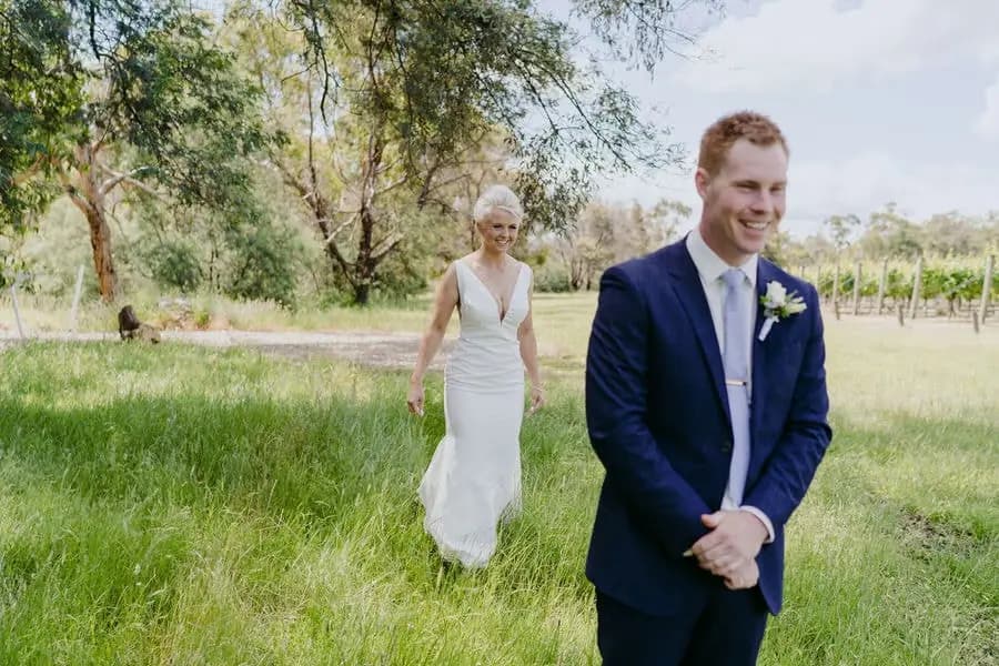 Groom smiles as the bride approaches behind him through long grass.