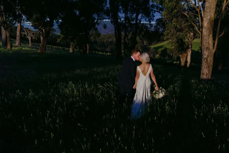 Couple embraces in a dark grassy field beneath trees at dusk.