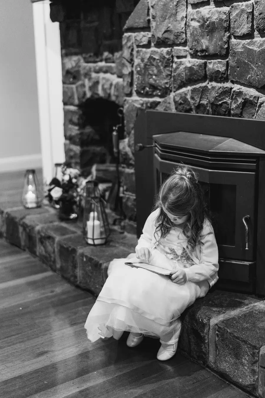 Black and white photograph of a small girl in a white dress seated beside a stone fireplace.
