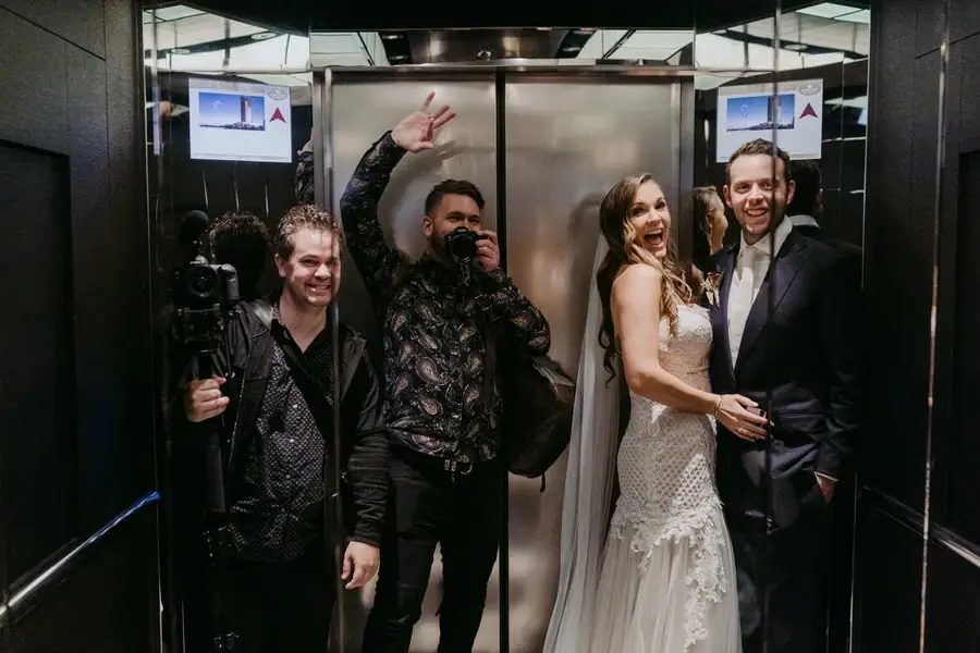Couple and guests smile inside a lift with a photographer holding a camera.