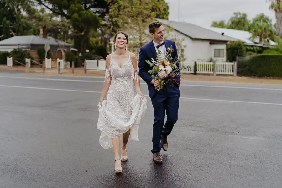 Couple walks across a quiet road with the groom carrying the bouquet.
