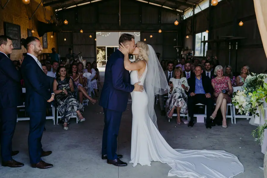 Couple kisses beneath the bride's veil as guests watch indoors.