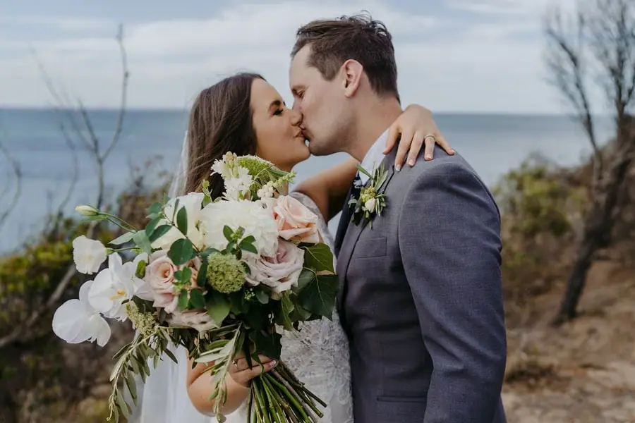 Couple kisses overlooking the sea with a large white bouquet in the foreground.