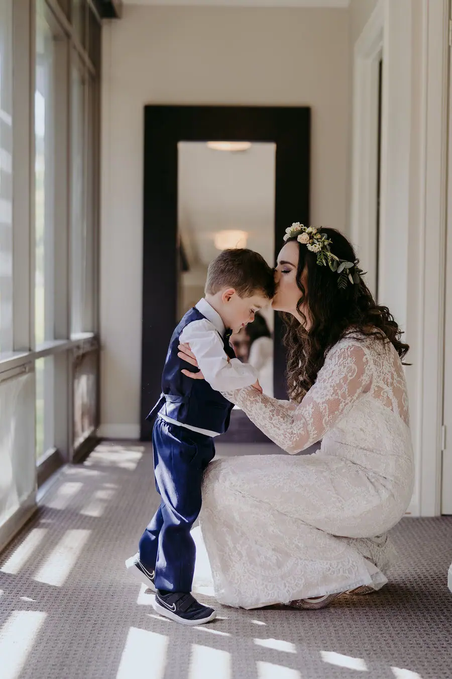 Bride kneels to embrace a small boy beside a window.