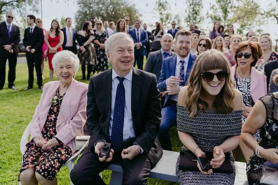 Seated ceremony guests laugh together on a garden lawn.
