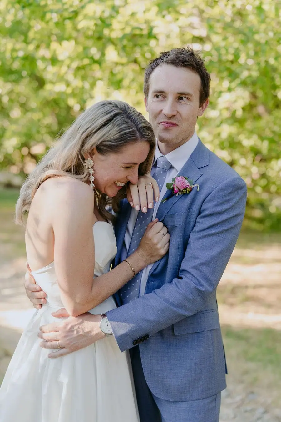 Bride rests her head against the groom as they embrace beneath a leafy tree.