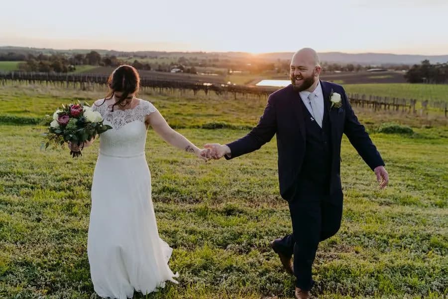 Couple holds hands and walks apart playfully on a lawn at sunset.