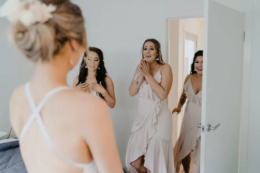 Bridesmaids react excitedly to the bride's dress indoors.