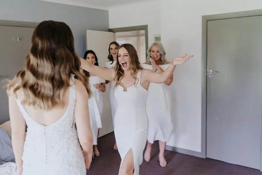 Bridesmaids open their arms in surprise toward the bride indoors.
