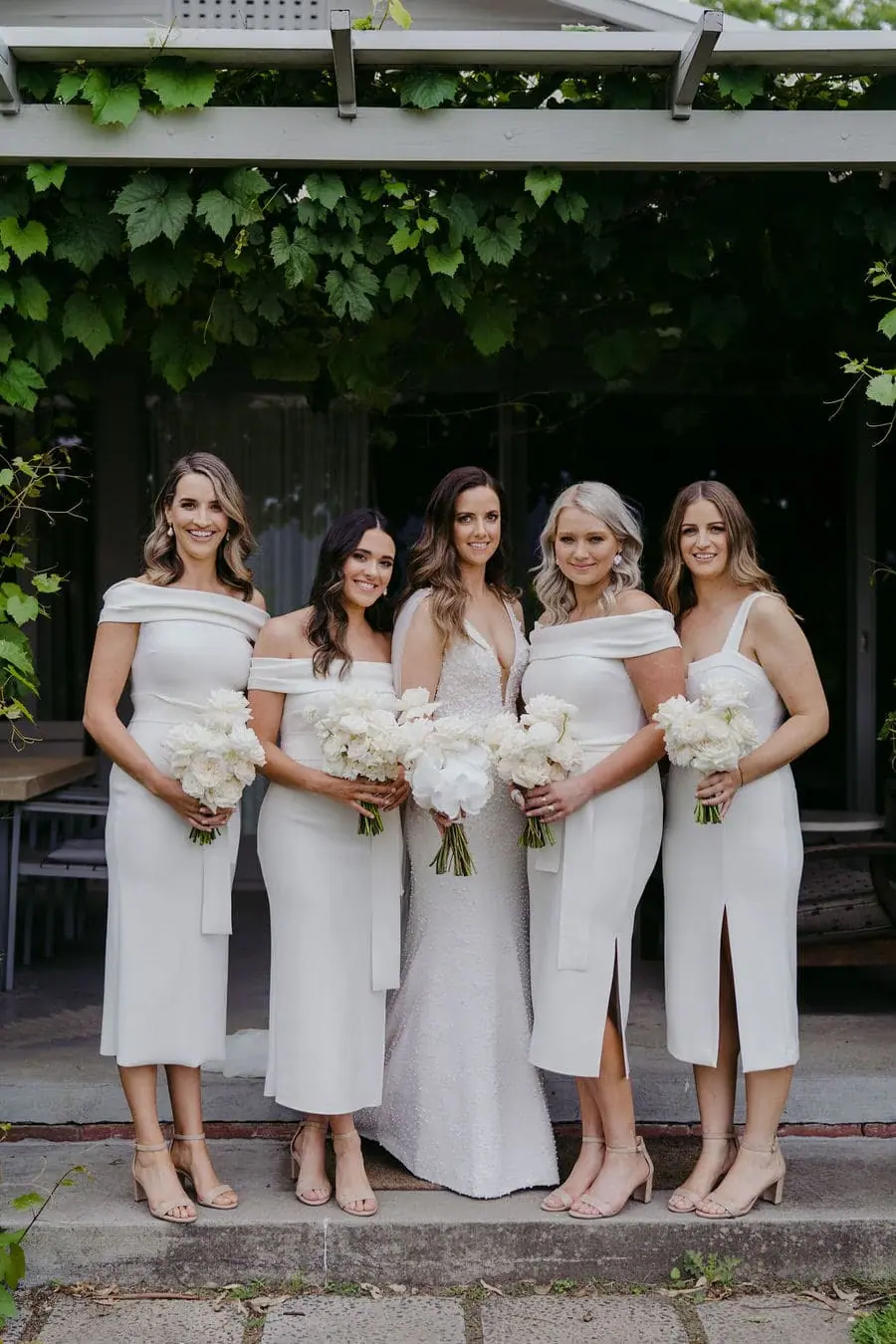 Bride and bridesmaids pose with bouquets beneath climbing greenery.