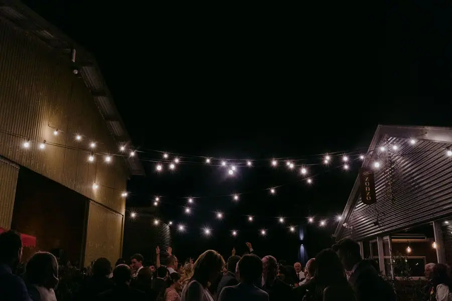 Crowd gathers beneath string lights between dark venue buildings at night.