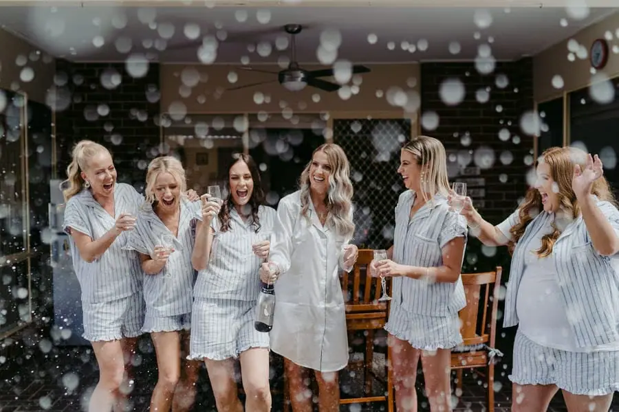 Women in striped pyjamas laugh as champagne sprays toward the camera.