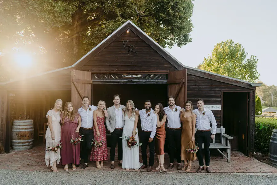 Wedding party poses outside a rustic wooden building in evening sunlight.