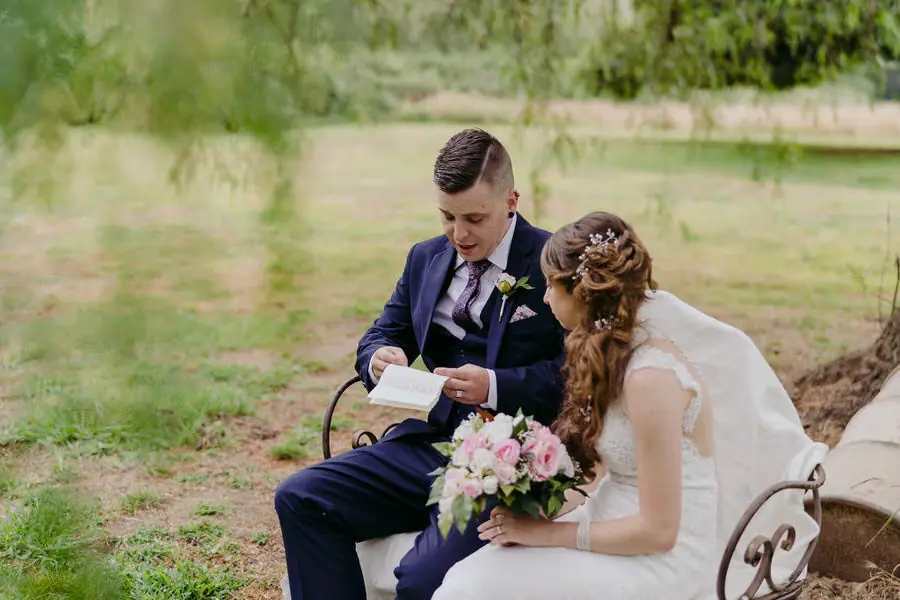 Seated groom opens a small box beside the bride in a garden.