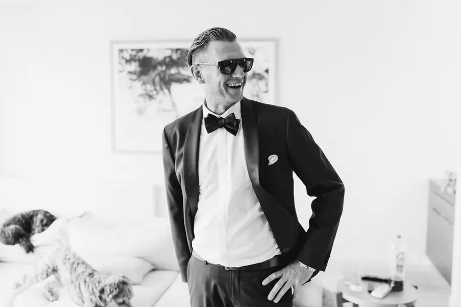 Black and white portrait of a smiling man in a tuxedo and sunglasses indoors.
