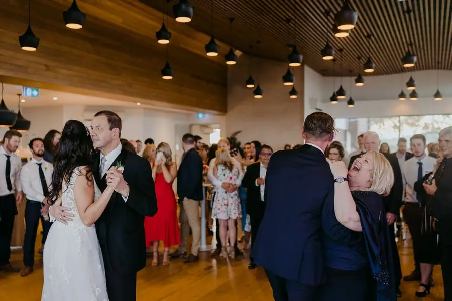 Two couples dance as guests watch in a bright reception room.