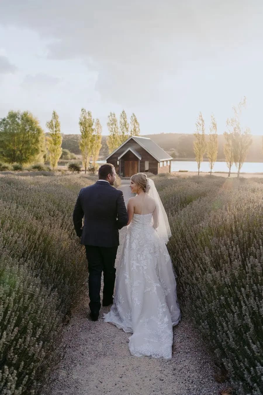 Rear view of the couple walking between rows of flowering plants toward a small building.