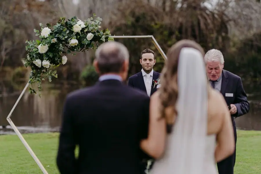 Groom waits beneath a geometric floral arch as the bride approaches with an older man.