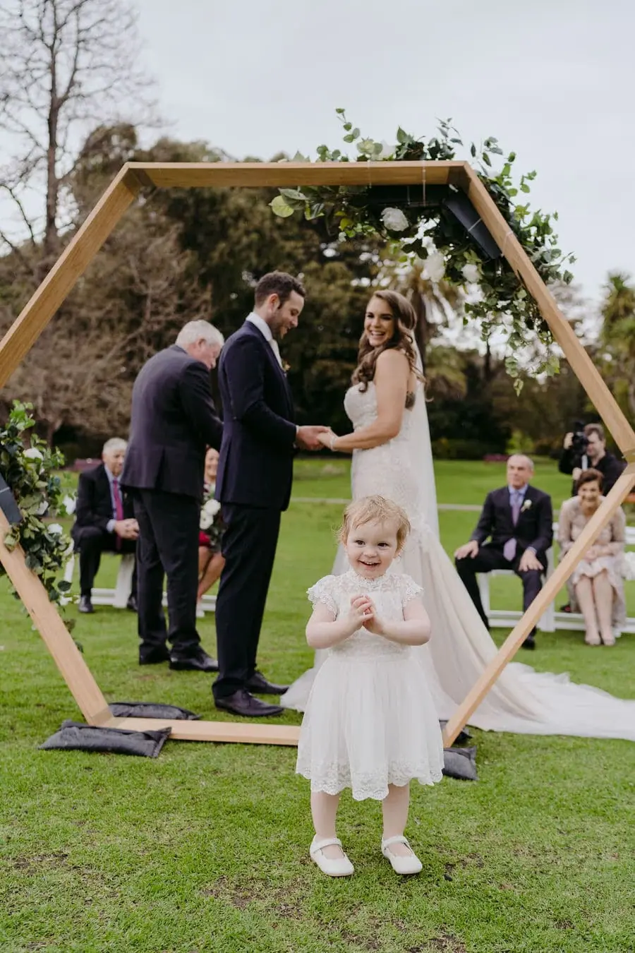 Small flower girl stands before a geometric arch as the couple holds hands behind her.