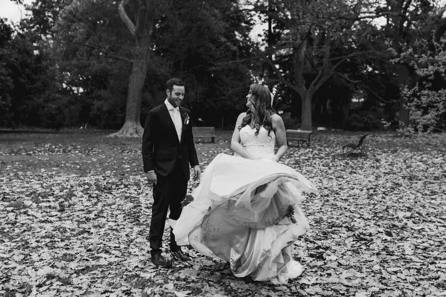 Black and white photograph of the couple walking across fallen leaves with the bride lifting her skirt.