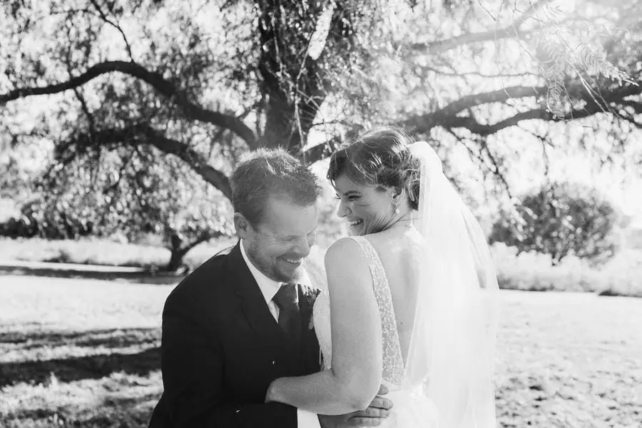 Black and white photograph of the groom embracing the bride beneath a sunlit tree.