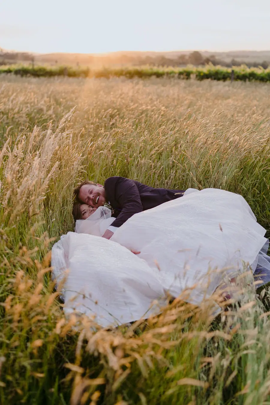Couple lies together in long grass with the bride's dress spread around them.
