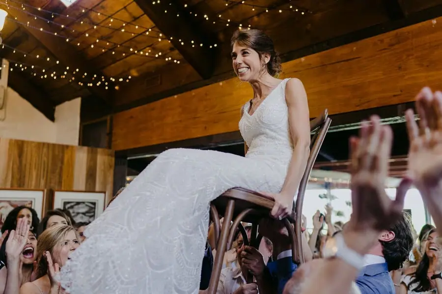 Guests lift the smiling bride in a chair beneath reception string lights.