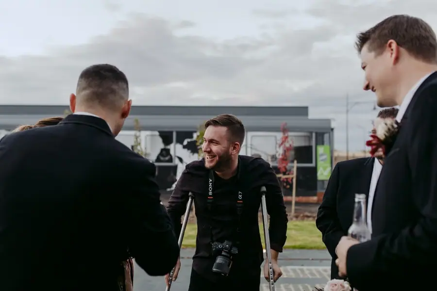 Photographer laughs with wedding guests outdoors while carrying a camera.