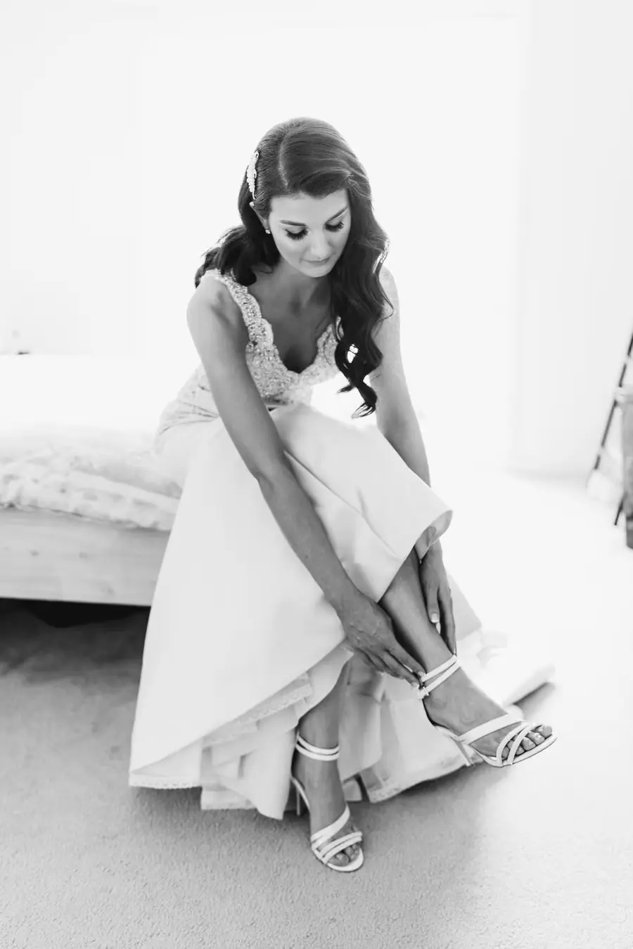 Black and white photograph of the bride fastening her shoe while seated indoors.