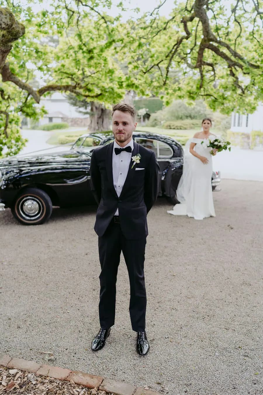 Groom waits in front of a vintage car as the bride approaches behind him.