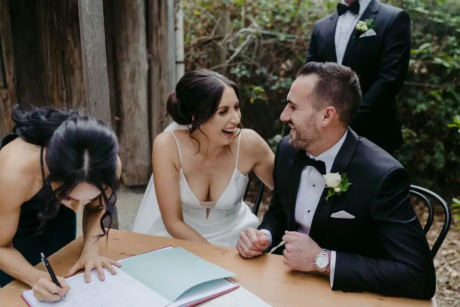 Couple laughs beside wedding paperwork as a woman signs at the table.