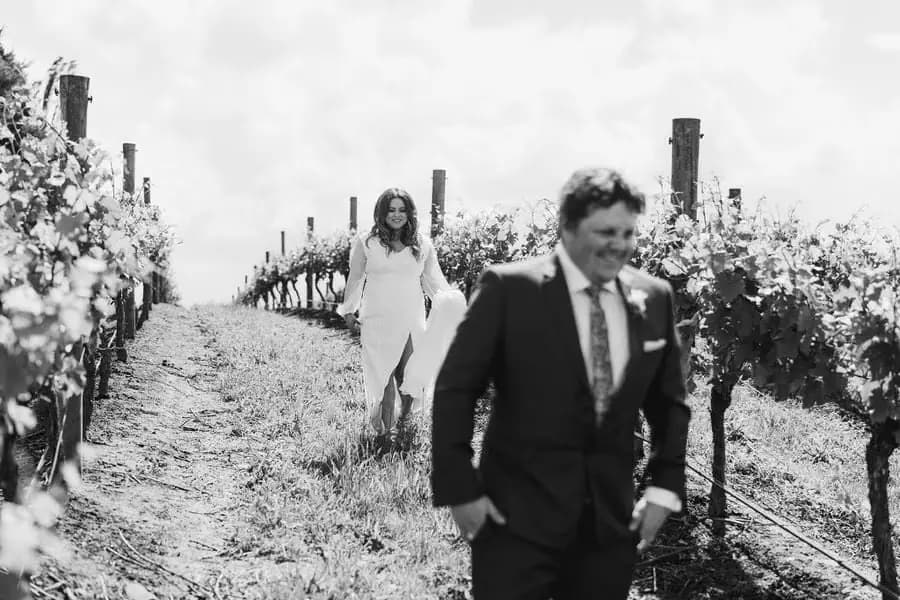 Black and white photograph of the bride approaching the groom between vineyard rows.