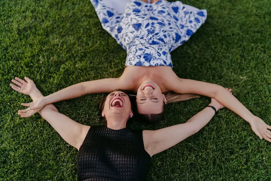 Two women lie on the grass laughing with their arms spread out.