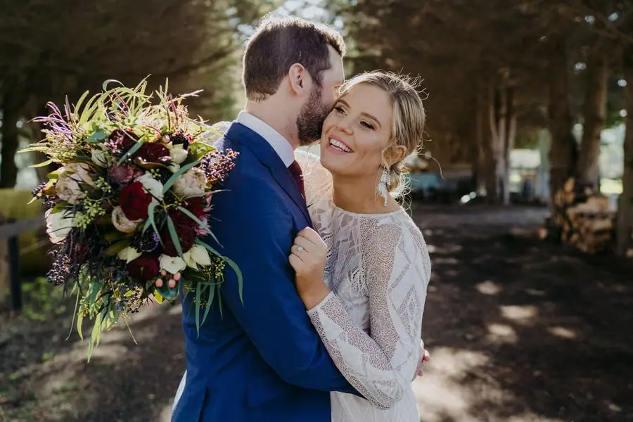 Groom kisses the bride's forehead with a colourful bouquet over his shoulder.