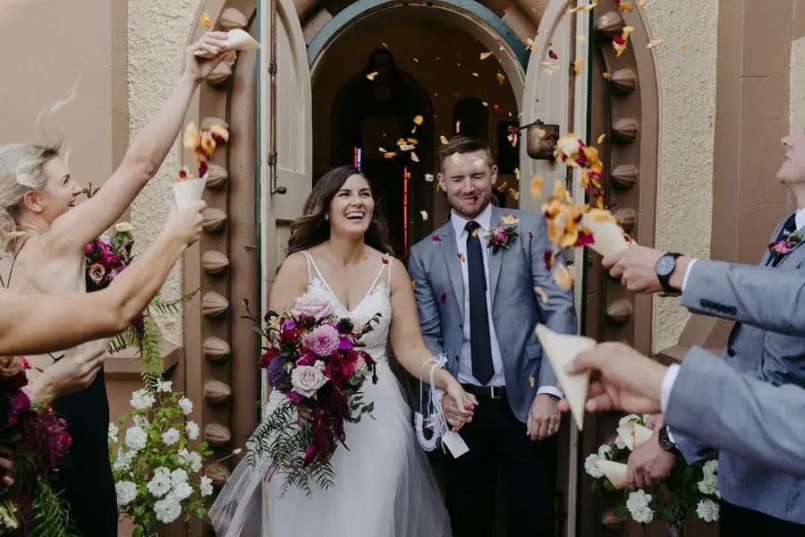 Newlyweds leave a church doorway through confetti thrown by guests.