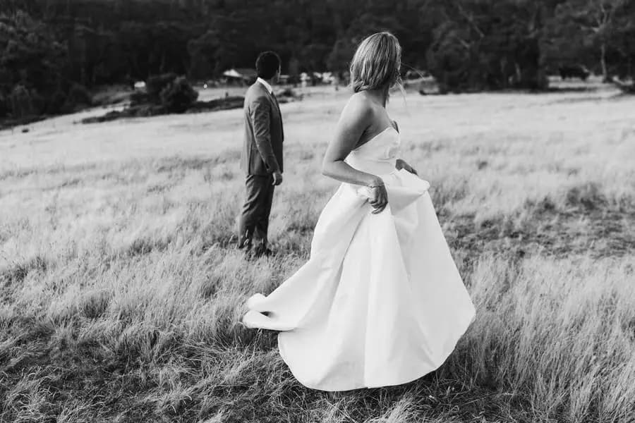 Black and white photograph of the bride gathering her dress in a grassy field.