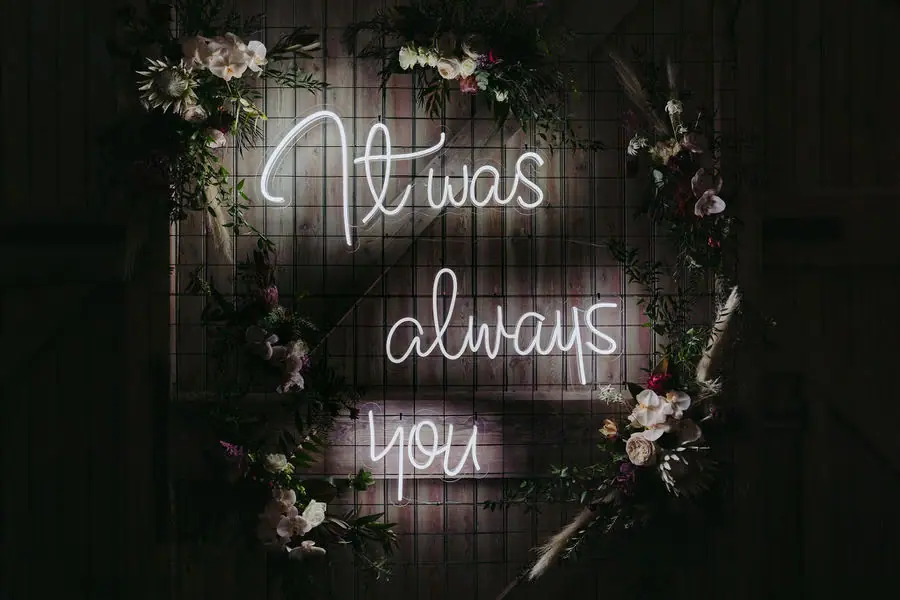 Glowing romantic sign surrounded by flowers against a dark reception wall.