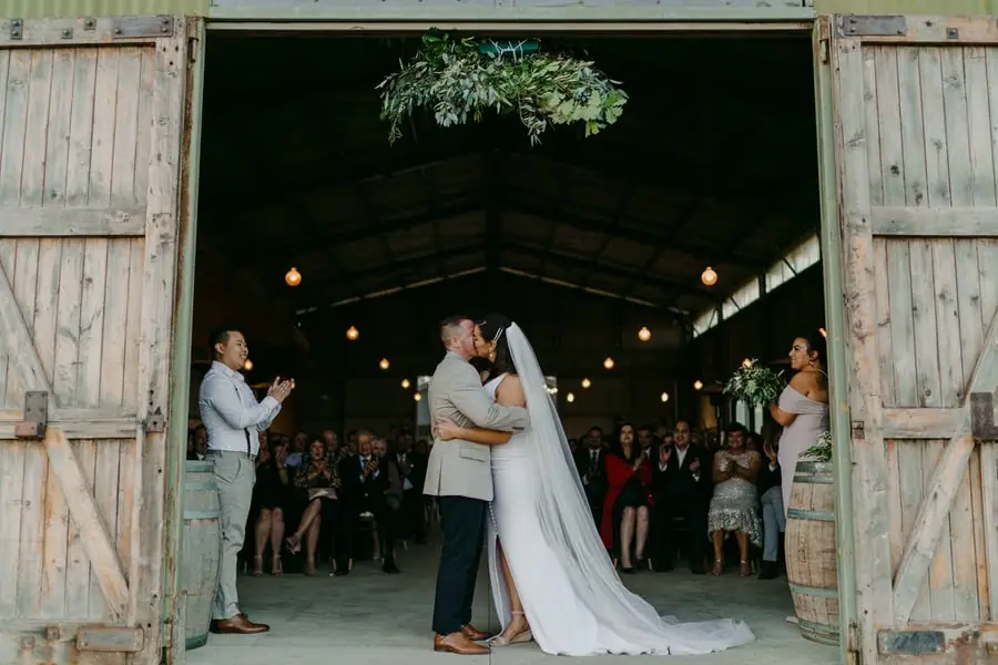 Couple kisses in a barn doorway with seated guests watching inside.