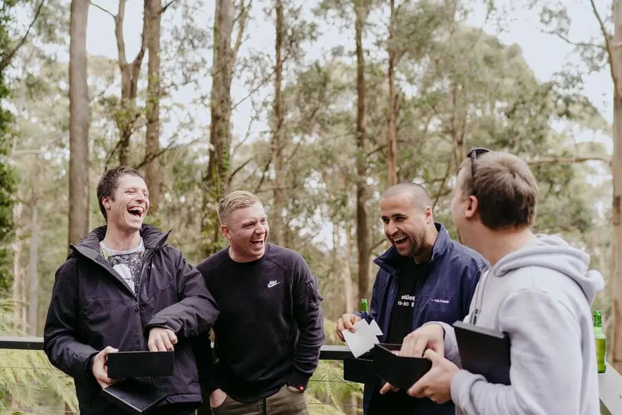 Men laugh together while opening boxes outdoors among trees.