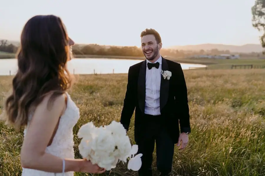 Groom smiles toward the bride in a grassy field beside water.