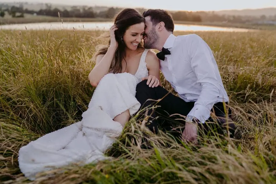 Couple sits close together in long grass beside water at sunset.