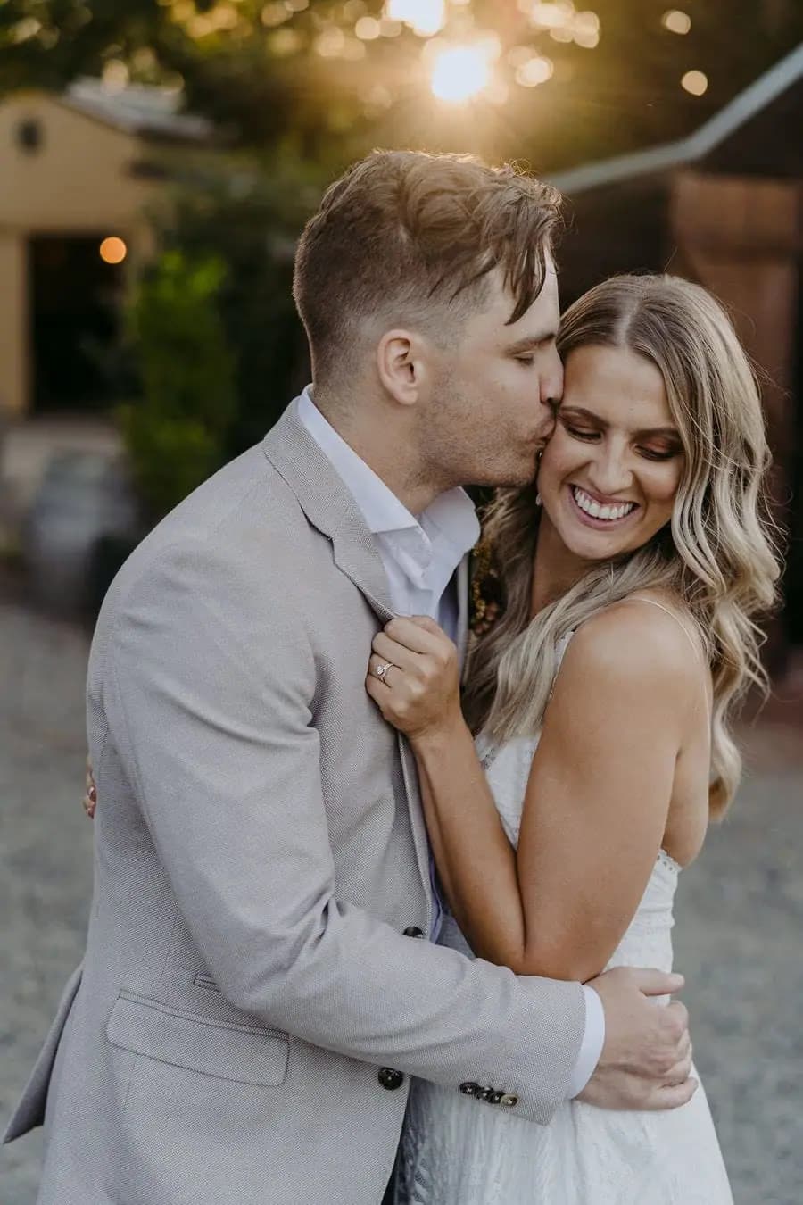Groom kisses the bride's forehead as they embrace in warm light.
