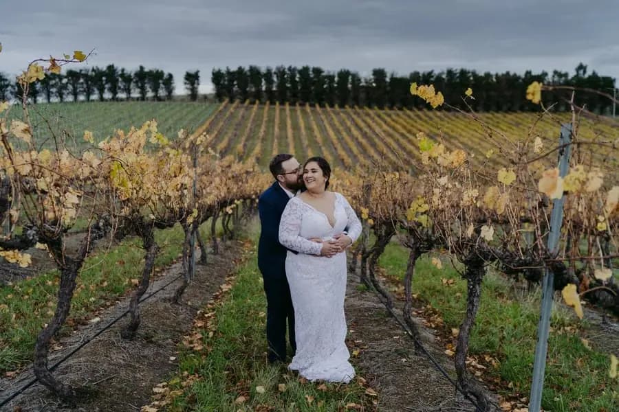 Groom embraces the bride from behind between autumn vineyard rows.