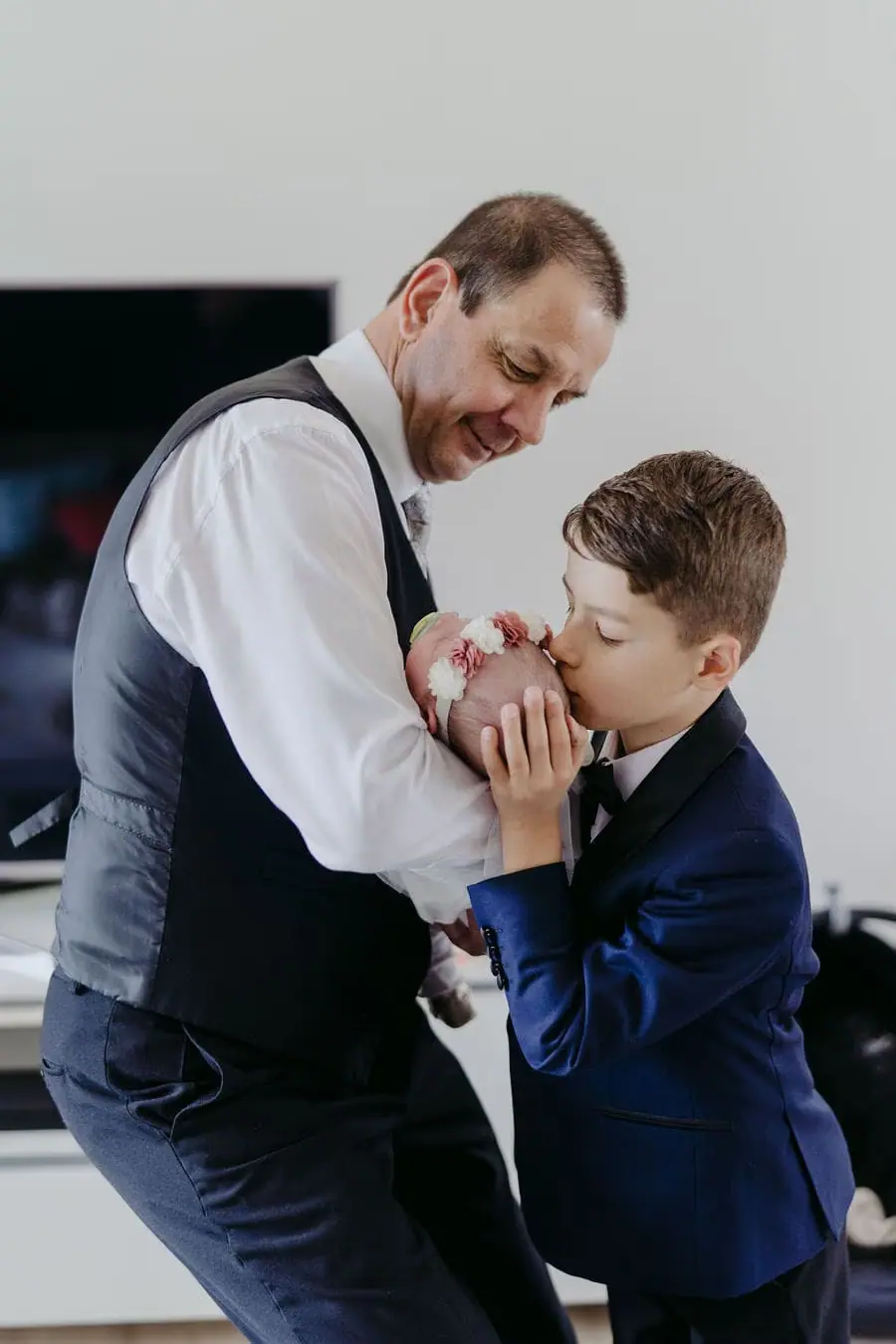 Man and a young boy in suits playfully clasp hands indoors.