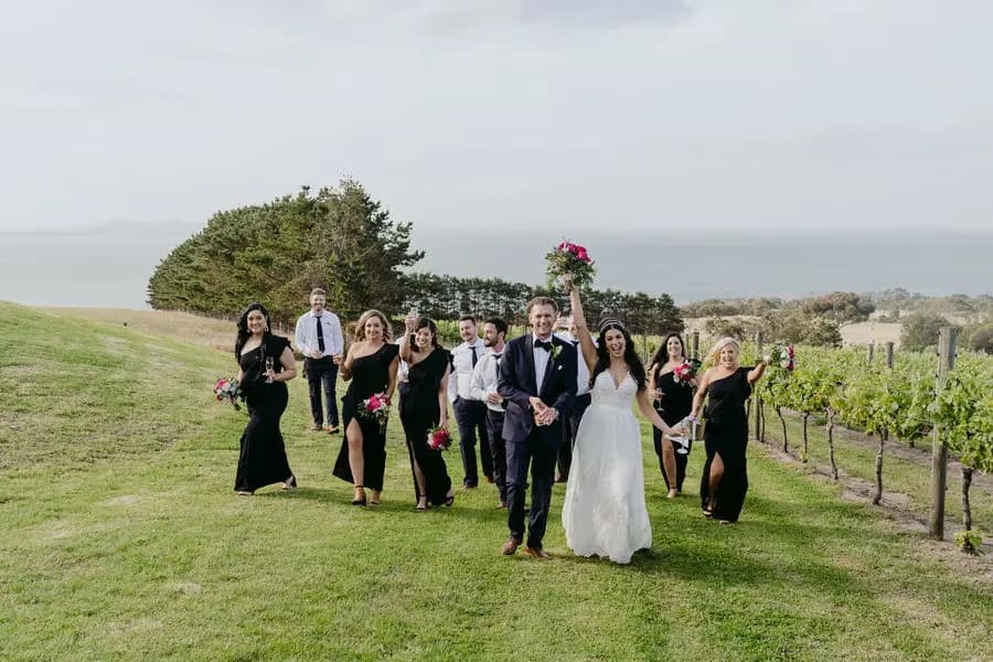 Wedding party walks across a grassy slope beside vineyard rows and the coast.