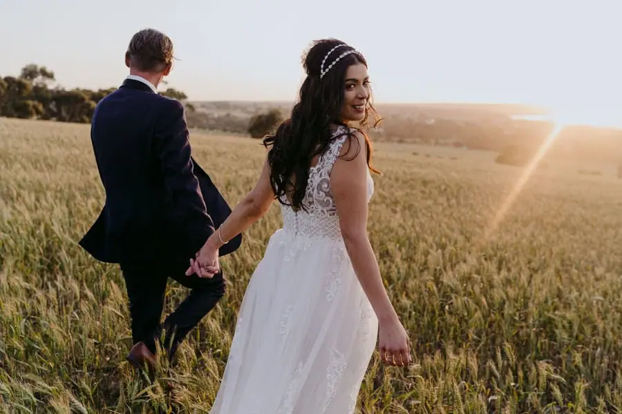 Couple walks hand in hand through a sunlit field as the bride looks back.