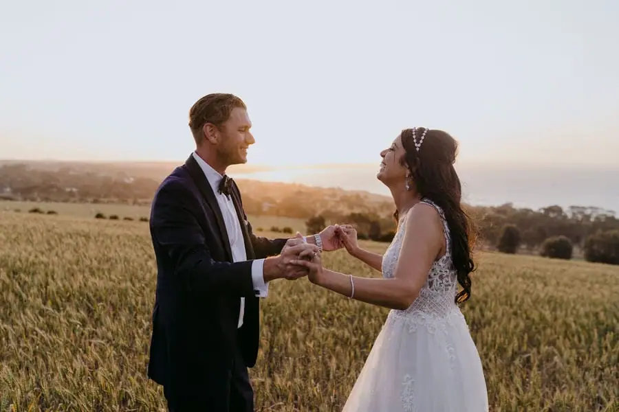 Couple holds both hands facing each other in a field at sunset.