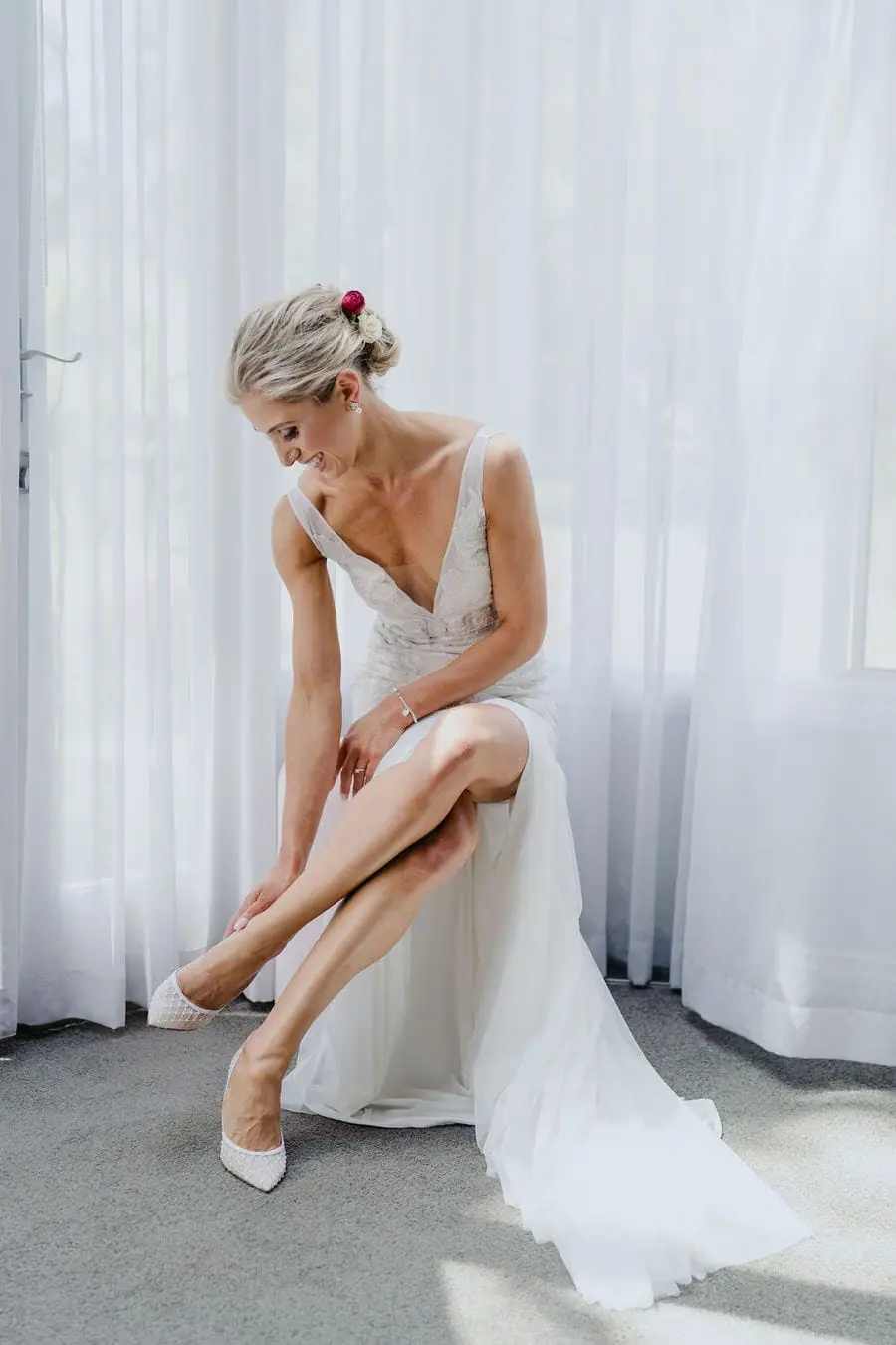 Bride sits beside a window fastening her shoe.