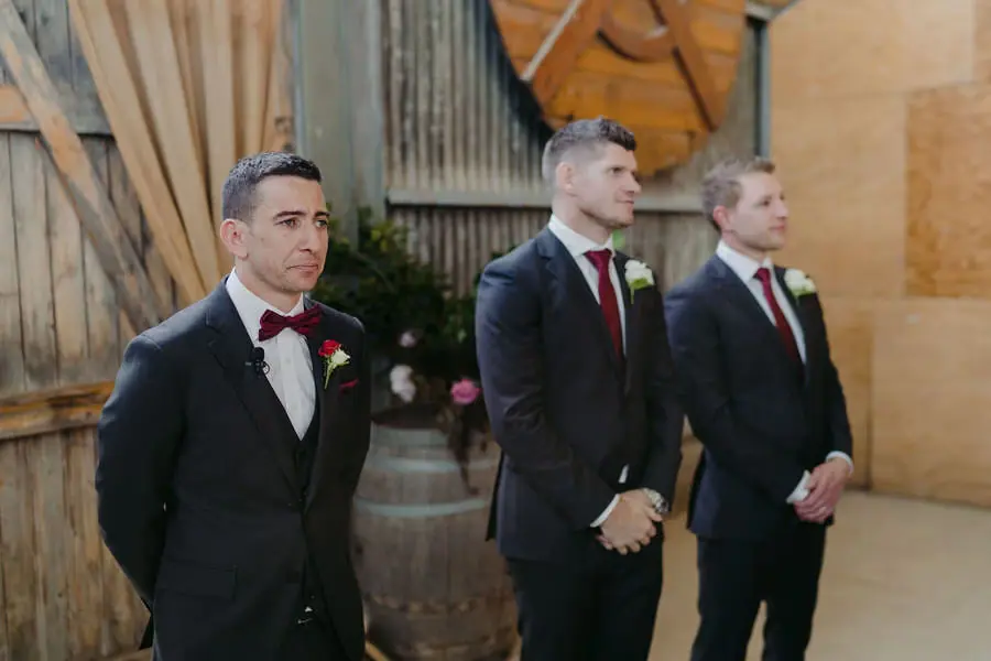 Groom and two groomsmen wait beside a wooden ceremony building.