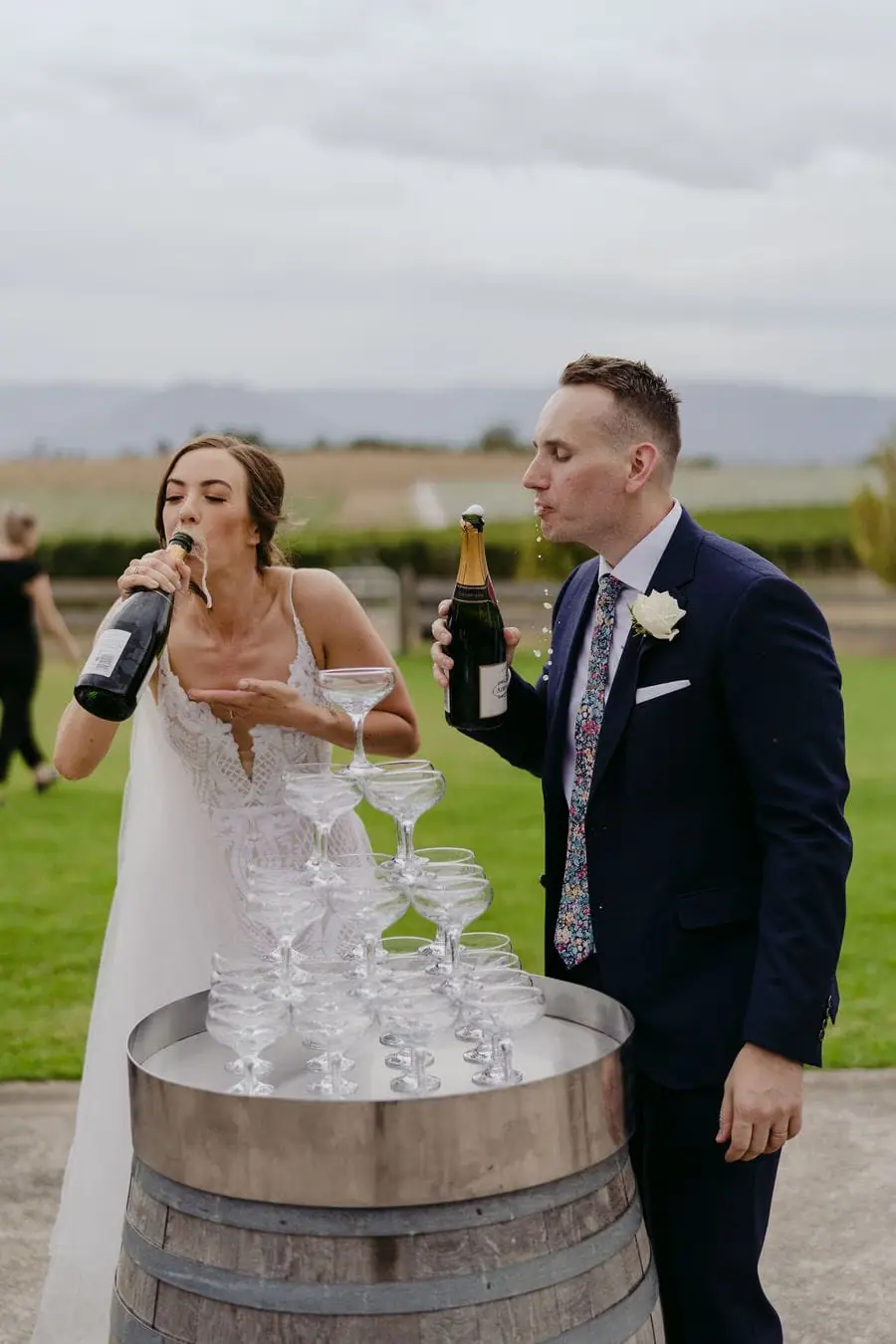 Couple drinks from bottles beside a tower of champagne glasses on a barrel.